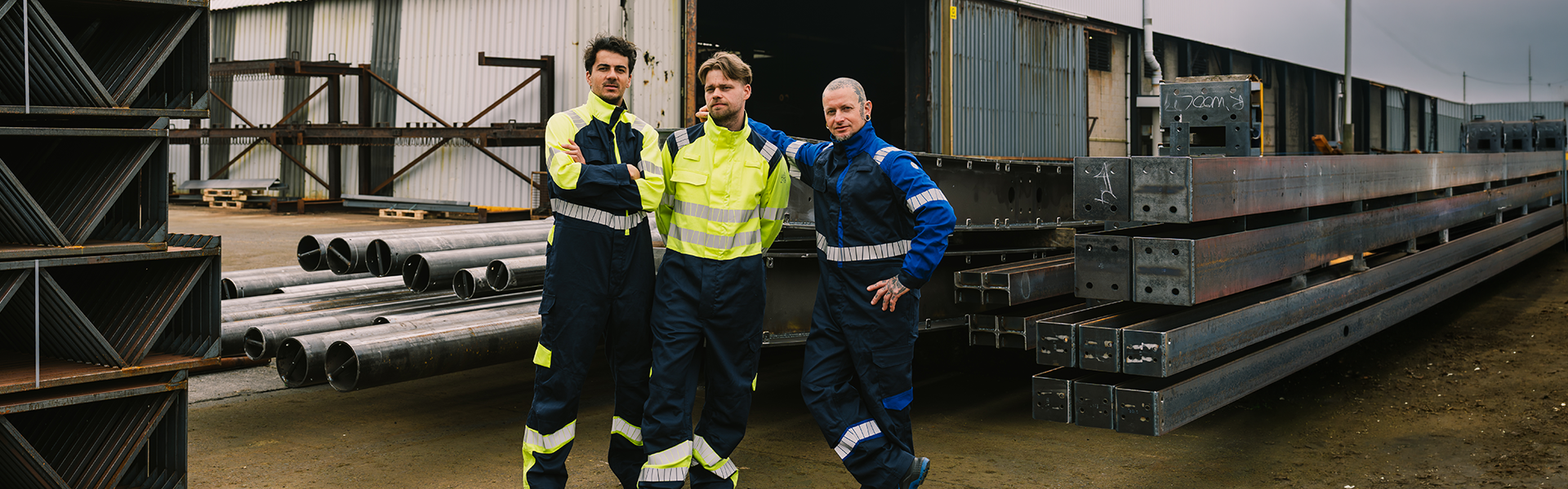 Drei Mitarbeiter tragen Warnschutz-Arbeitskleidung auf einem Industriegelände vor gelagerten Stahlträgern und Metallrohren.