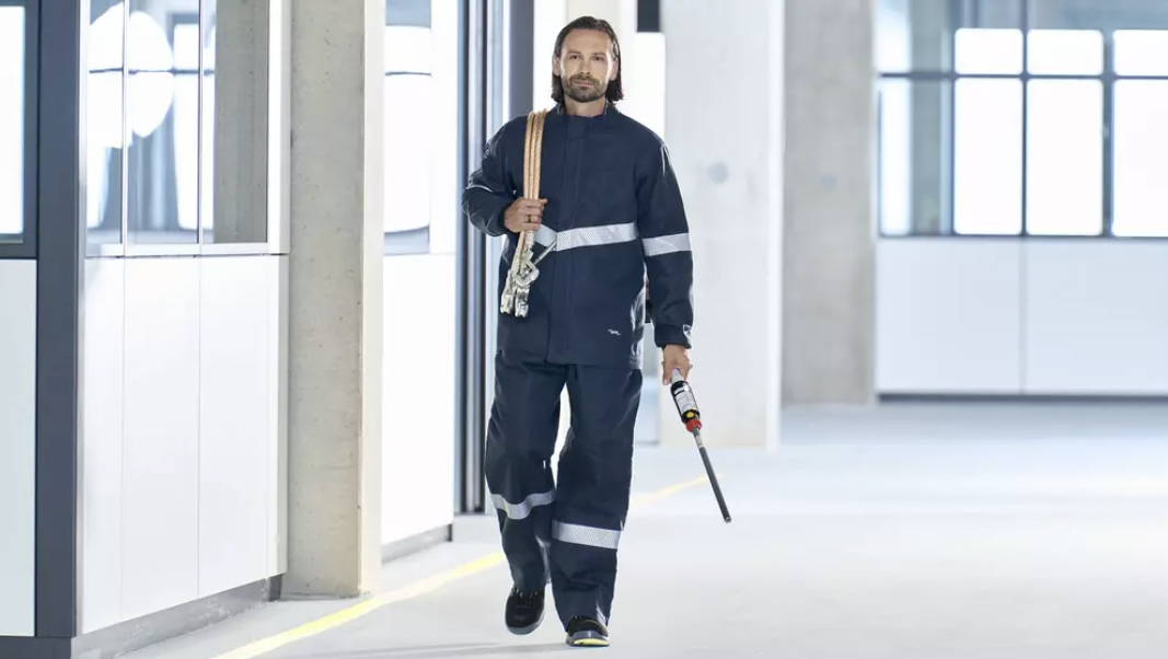 Mann in dunkler Arbeitskleidung mit Reflexstreifen trägt Seil und Werkzeug in einer Industriehalle