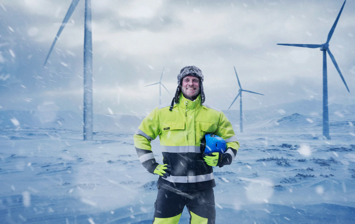 Arbeiter in gelb-schwarzer Warnschutz-Winterjacke und Handschuhen vor Windkraftanlagen im Schneesturm mit Schutzhelm in der Hand