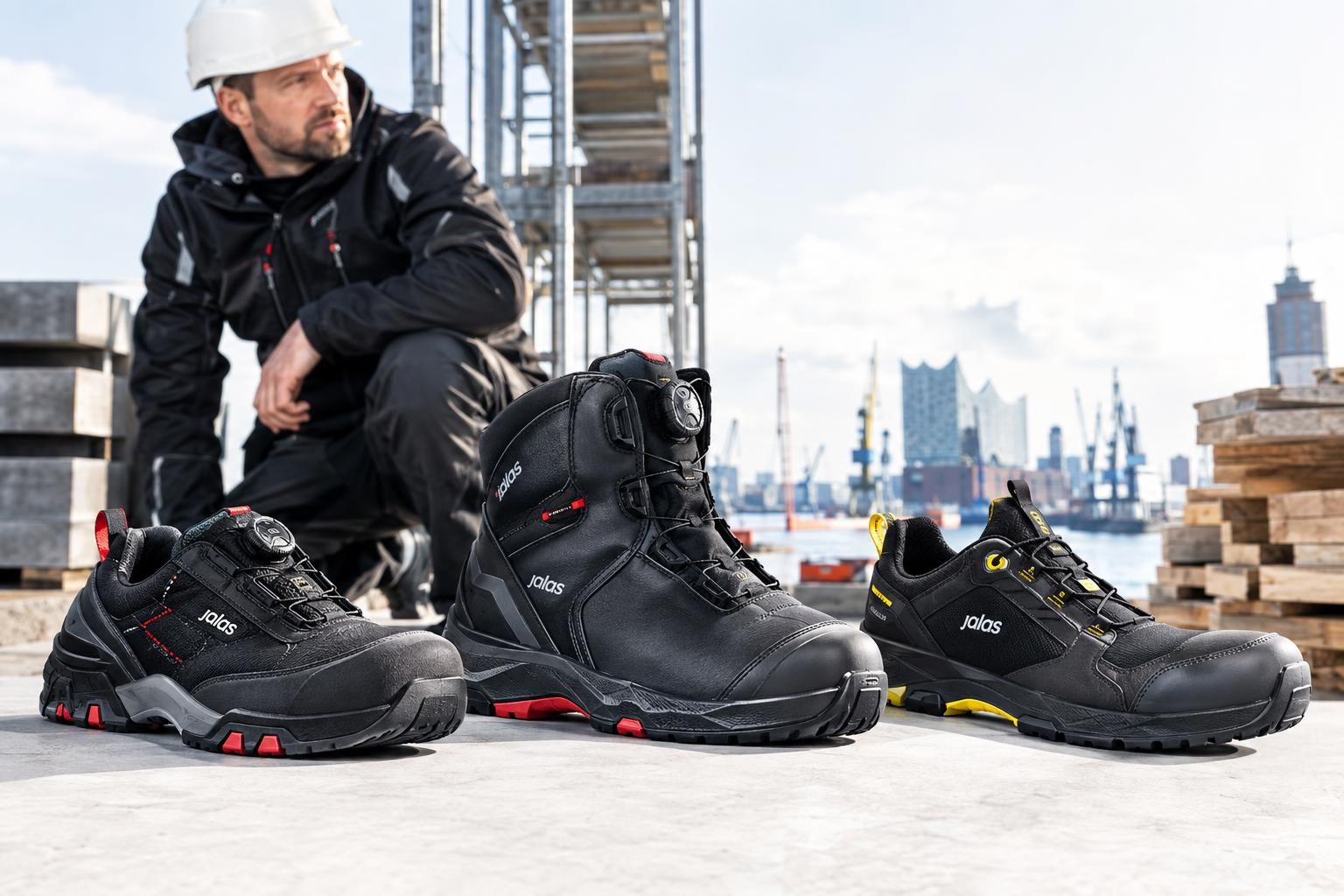 Drei schwarze JALAS Sicherheitsschuhe und ein Sicherheitsstiefel auf einer Baustelle, daneben ein Arbeiter mit Helm vor Industriehafen-Kulisse