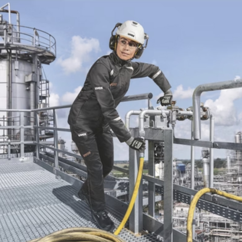 Frau im Industrieeinsatz trägt die Uvex suXXeed Arc Damen MultiNorm Jacke 4 kA + 7 kA APC2 von Uvex auf einer Anlage mit Rohrleitungen.
